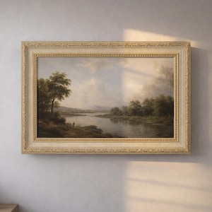 Puede incluir: Una pintura de paisaje enmarcada representa una serena escena fluvial con árboles y una figura en la orilla. La obra de arte está encerrada en un marco decorativo de color crema, que añade un toque de elegancia. La pintura evoca una sensación de tranquilidad.