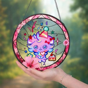 Może przedstawiać: Okrągłe witrażowe okno z postacią z kreskówki w czapce imprezowej, otoczone świątecznymi dekoracjami. Okno ma różową kwiatową obwódkę i jest zawieszone na łańcuchu. Postać ma twarz w kształcie serca.