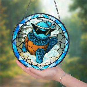 Może przedstawiać: Okrągłe witrażowe dzieło sztuki przedstawiające kreskówkowego żółwia w okularach przeciwsłonecznych i niebieskiej bandanie. Żółw ma niebieski korpus i pomarańczowy pancerz. Grafika jest oprawiona w czarną ramę i łańcuch do zawieszenia.