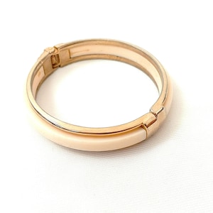 May include: A hinged bangle bracelet featuring a polished gold-tone metal frame and a smooth, cream-colored inlay. The circular accessory includes a secure clasp mechanism on one side, showcasing a clean, metallic finish and a neutral color palette.