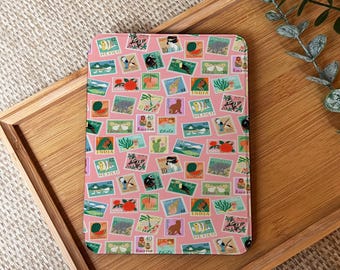 May include: A rectangular pink notebook cover adorned with a collage of colorful postage stamps. The stamps feature various illustrations, including animals, fruits, and landscapes, with text such as "MEXICO" and "INDIA". The notebook is placed on a wooden tray.