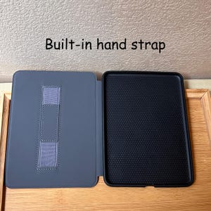 Puede incluir: Funda de tableta gris abierta con una correa de mano incorporada. El interior es negro con un patr&oacute;n texturizado. El texto "Built-in hand strap" est&aacute; encima de la funda.