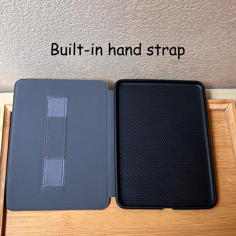Puede incluir: Funda de tableta gris abierta con una correa de mano incorporada. El interior es negro con un patr&oacute;n texturizado. El texto "Built-in hand strap" est&aacute; encima de la funda.