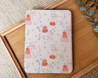 Puede incluir: Un objeto decorativo rectangular de color rosa claro con temática de gatos. El diseño presenta dibujos lineales de gatos con lazos, calabazas y estrellas. Las calabazas son naranjas y rosas. El artículo está sobre una bandeja de madera.