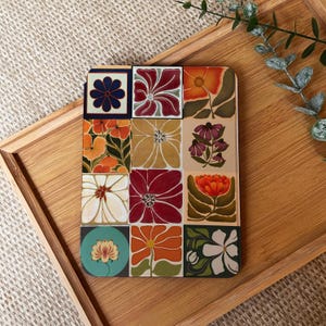 May include: A square coaster with a floral pattern. The coaster is divided into a grid of squares, each displaying a different flower in shades of red, orange, yellow, and blue. It rests on a wooden tray.