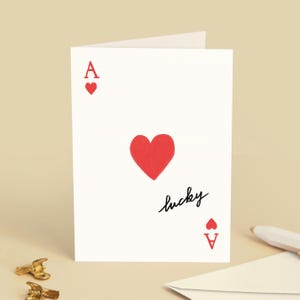 Könnte beinhalten: Weiße Grußkarte mit einem Herz-Ass-Design. Die Karte zeigt ein großes rotes Herz, mit kleineren roten Herzen und dem Buchstaben "A". Das Wort "lucky" ist in Schreibschrift geschrieben. Schlichtes und elegantes Design.