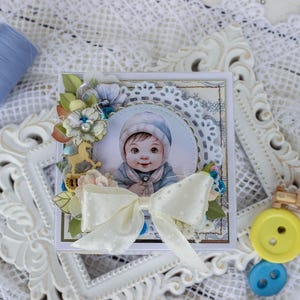 Puede incluir: Una caja decorativa cuadrada con una imagen circular central de un bebé con gorro azul, adornada con un gran lazo de cinta color crema con lunares, flores de papel y un pequeño dije de madera de caballito balancín sobre un marco de encaje.