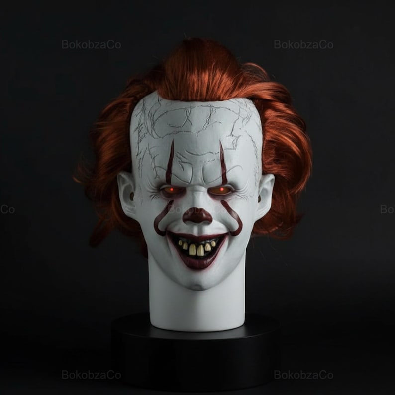 May include: A white clown mask with red hair, red eyes, and a sinister grin. The mask features red markings around the eyes and mouth, and is mounted on a black base. The mask is a prop for Halloween or cosplay.