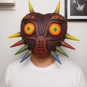 May include: A detailed, burgundy mask with yellow, red, green, and blue spikes. The mask features large, orange eyes with green pupils and intricate detailing. The mask is worn by a person wearing a white t-shirt.