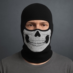 May include: Black balaclava featuring a skull design. The skull is rendered in white and grey, with detailed teeth and eye sockets. The balaclava covers the head and neck, with only the eyes exposed.