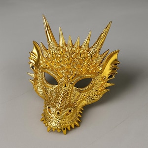May include: A gold dragon mask with detailed features. The mask has spikes, eye openings, and a textured surface. It is designed for wear, possibly for a costume or masquerade. The mask's design is reminiscent of a mythical creature.