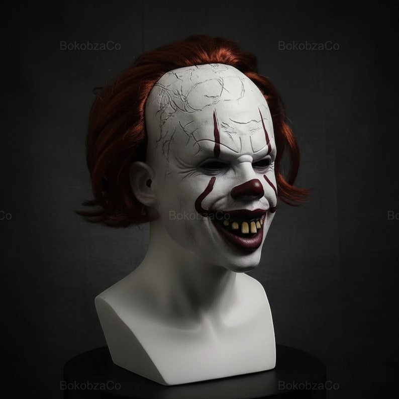 May include: A white clown mask with red accents, including a nose, mouth, and facial markings. The mask has a cracked texture on the forehead and a sinister grin revealing yellow teeth. The mask is set on a white bust with red hair.