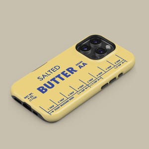 May include: A yellow phone case with a stick of butter design. The case has a black border and a blue label that reads "SALTED BUTTER GRADE AA". The label also includes a measurement guide in tablespoons and cups.