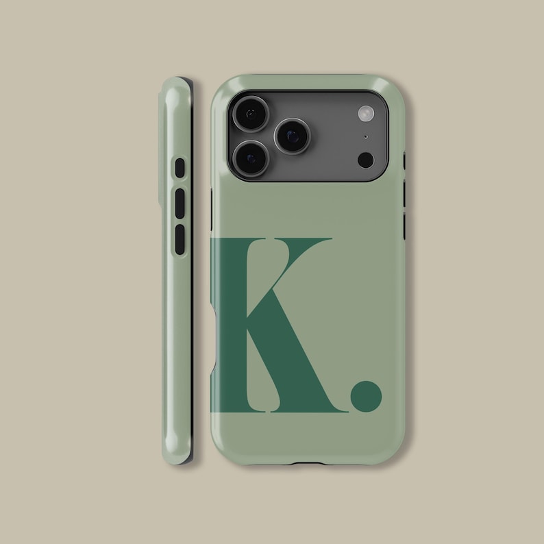 Op de afbeelding: Saliegroene telefoonhoes met een donkergroene letter "K" en een punt. De hoes heeft een camera-uitsparing en zijknoppen. De telefoonhoes wordt van voren en van de zijkant getoond.