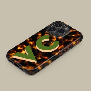 Puede incluir: Una funda de tel&eacute;fono negra con un patr&oacute;n de caparaz&oacute;n de tortuga y las letras "VC" en verde y beige.