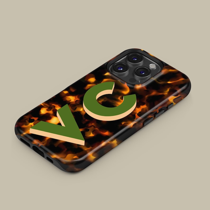 Puede incluir: Una funda de tel&eacute;fono negra con un patr&oacute;n de caparaz&oacute;n de tortuga y las letras "VC" en verde y beige.