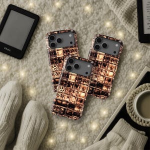 Puede incluir: Tres fundas para teléfono con un diseño de mosaico marrón y dorado se muestran sobre una superficie suave y blanca texturizada. Las fundas tienen una carcasa de cámara gris oscuro. También se ven un lector de libros electrónicos negro y una taza de café.
