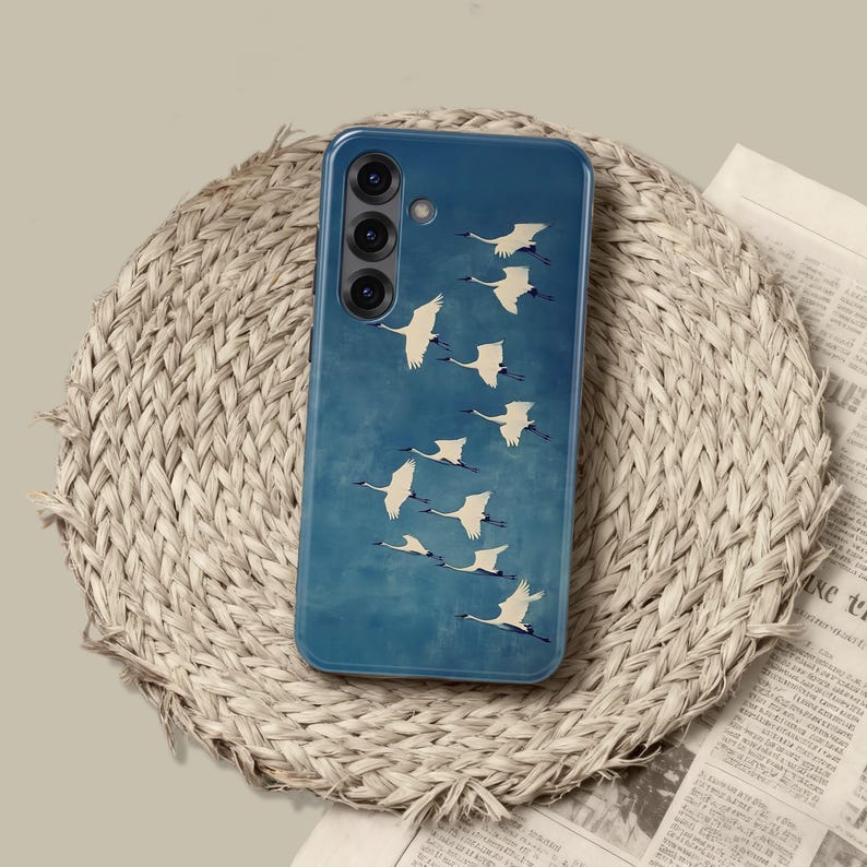 May include: A blue phone case featuring a flock of white cranes in flight against a textured background. The case has a clear, protective cover and a camera array on the upper left. The case rests on a woven, circular placemat.
