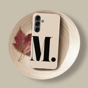 Op de afbeelding: Een beige telefoonhoesje met de letter "M" in een vetgedrukte zwarte letter. De telefoon ligt op een houten bord met een rood blad. De telefoon heeft een drievoudige cameralens. Het bord heeft een diameter van ongeveer 20 cm.