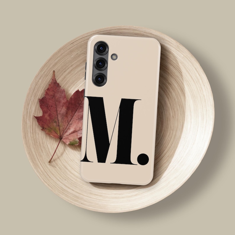 Op de afbeelding: Een beige telefoonhoesje met de letter "M" in een vetgedrukte zwarte letter. De telefoon ligt op een houten bord met een rood blad. De telefoon heeft een drievoudige cameralens. Het bord heeft een diameter van ongeveer 20 cm.