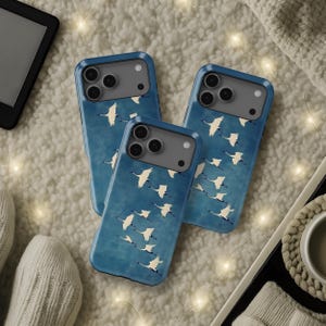 May include: Three blue phone cases with a white crane design. The cases have a matte finish and a raised camera bump. The background is a soft, textured surface with warm lighting.