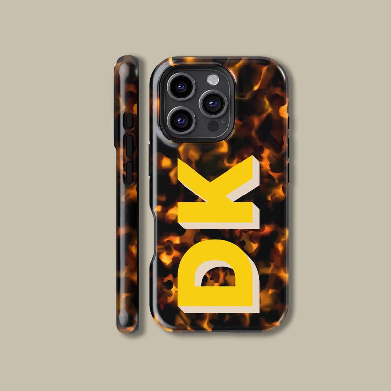 Puede incluir: Una funda de tel&eacute;fono negra con un patr&oacute;n de caparaz&oacute;n de tortuga y las letras "DK" en amarillo. La funda tiene un recorte para el objetivo de la c&aacute;mara y un recorte para el bot&oacute;n de volumen.