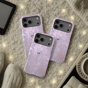 Op de afbeelding: Drie telefoonhoesjes met een roze glas-in-lood ontwerp met een spookillustratie. De hoesjes liggen op een zacht, getextureerd oppervlak. De hoesjes hebben een zwarte cameramodule.
