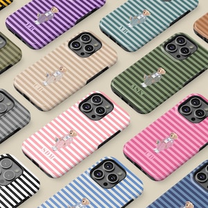 Op de afbeelding: Een verzameling telefoonhoesjes met een gestreept patroon in verschillende kleuren, waaronder roze, blauw, groen en beige. Elke hoes heeft een cartoon beer illustratie en initialen. De hoesjes zijn voor een smartphone.