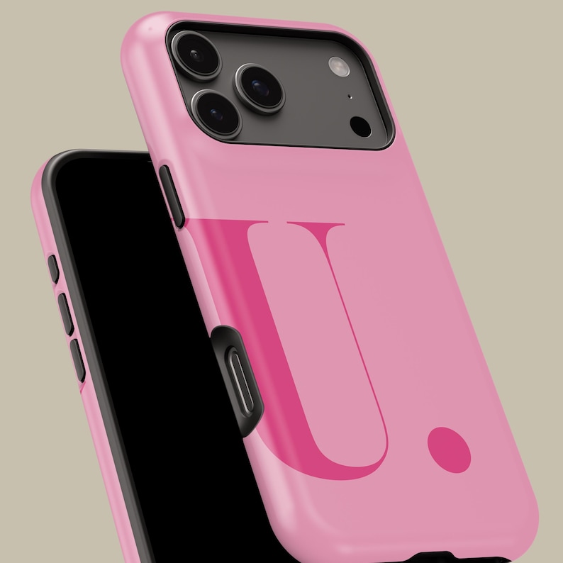 Op de afbeelding: Een roze telefoonhoesje met de letter "U" in een donkerdere roze tint. De hoes is schuin geplaatst, waardoor het zwarte scherm van de telefoon en de cameralenzen zichtbaar zijn. Onder de letter staat een roze stip.
