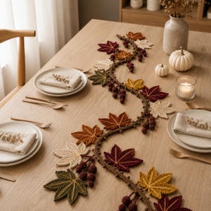 Puede incluir: Un camino de mesa de ganchillo que presenta una enredadera sinuosa con bayas de color marrón oscuro y varias hojas de colores otoñales en tonos burdeos, amarillo mostaza, verde oliva y crema, dispuestas en un patrón decorativo sobre una mesa de madera.