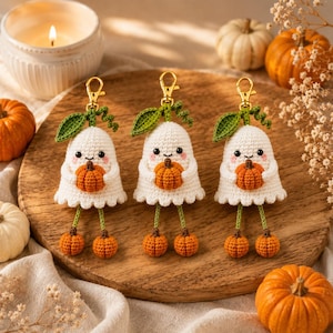 Puede incluir: Tres llaveros de fantasmas tejidos a ganchillo en color blanco, cada uno sosteniendo una pequeña calabaza naranja. Los fantasmas tienen hojas verdes tejidas arriba, piernas colgantes con pies de calabaza y cierres de mosquetón dorados.