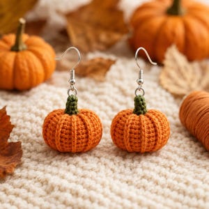 May include: A pair of dangle earrings featuring small, crocheted orange pumpkins with green stems. Each pumpkin is attached to a silver-toned metal fishhook ear wire, creating a textured, spherical design that hangs vertically from the hook.