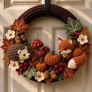 Op de afbeelding: Een ronde gehaakte krans met een donkerbruine basis, versierd met herfstthema-elementen waaronder een egel, een vos, twee pompoenen, rood-witte paddenstoelen, eikels, bessen en diverse esdoornbladeren in groen-, oranje- en roodtinten.
