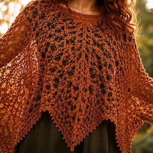 Op de afbeelding: Een roestkleurige gehaakte poncho met een ingewikkeld opengewerkt kantpatroon, cirkelvormige motieven aan de zijkanten en een puntige zoom. Het kledingstuk wordt over een donkerkleurige top gedragen en toont een gedetailleerd, luchtig breipatroon.