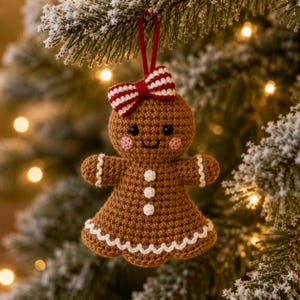 May include: A brown crocheted gingerbread person ornament featuring a red and white striped bow on its head, black bead eyes, pink stitched cheeks, and three white buttons on its chest. The edges are trimmed with white scalloped stitching.