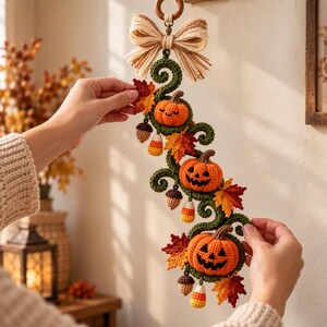 May include: A crocheted wall hanging featuring three orange pumpkins, two with jack-o'-lantern faces, accented by green vines, red and orange autumn leaves, small brown acorns, and candy corn shapes, topped with a beige fabric bow and wooden ring.