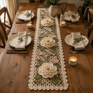 May include: A long, rectangular crochet table runner featuring an intricate cream-colored lattice pattern. The runner is decorated with three large, dimensional white crocheted flowers, smaller pink blossoms, and green crocheted leaves.