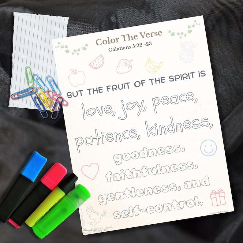 May include: A white coloring page with the text "Color The Verse" and "Galatians 5:22-23". The text "BUT THE FRUIT OF THE SPIRIT IS" is above the words "love, joy, peace, patience, kindness, goodness, faithfulness, gentleness, and self-control". Colorful highlighters and paperclips are also present.