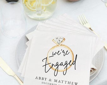 May include: White paper napkins with a gold ring design and the words "we're Engaged". The napkins also include the names "Abby & Matthew" and the date "December 28, 2024". The napkins are stacked on a plate.