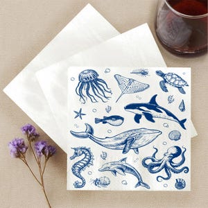 May include: White square paper napkins with a blue nautical design featuring various sea creatures, including a jellyfish, whale, dolphin, and octopus. The napkins are placed on a beige surface with a glass of red liquid in the background.