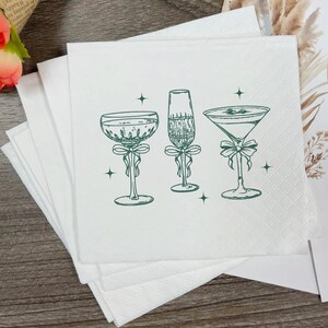 May include: White cocktail napkins with a green illustration of three different cocktail glasses, each adorned with a bow. The napkins are stacked, with a few visible in the foreground.