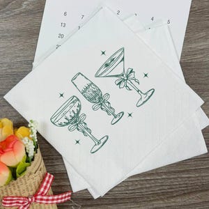 May include: White cocktail napkins with a green illustration of three different cocktail glasses, each adorned with a bow. The napkins are stacked on a wooden surface, with a calendar page visible in the background.