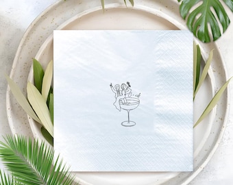 May include: A white square napkin with a black line drawing of two people in a cocktail glass. The napkin is on a white plate, surrounded by green leaves and a large green leaf.