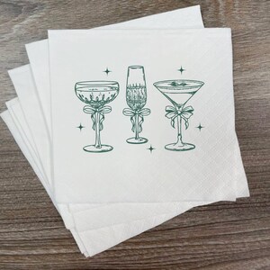 May include: White cocktail napkins with a green illustration of three different cocktail glasses, each adorned with a bow. The glasses include a champagne flute, a martini glass, and a coupe glass. Small star accents are scattered around the glasses.