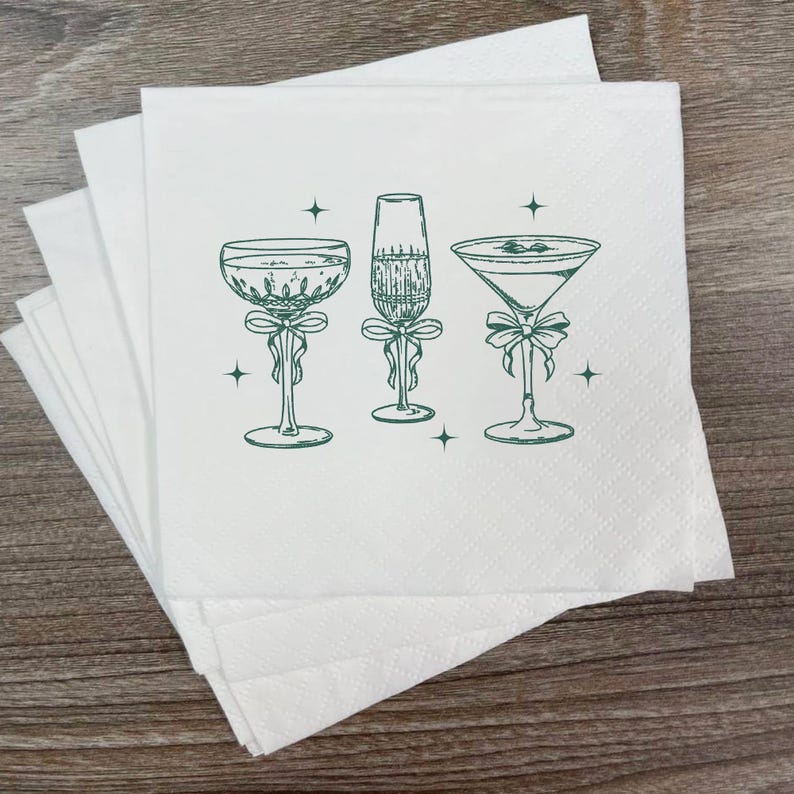 May include: White cocktail napkins with a green illustration of three different cocktail glasses, each adorned with a bow. The glasses include a champagne flute, a martini glass, and a coupe glass. Small star accents are scattered around the glasses.