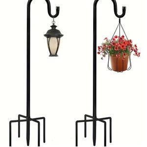 Pode incluir: Dois suportes de plantas de metal preto com um gancho curvo na parte superior. O suporte à esquerda tem uma lanterna pendurada no gancho. O suporte à direita tem um vaso pendurado com flores vermelhas.