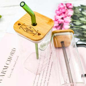 Może przedstawiać: Dwa przezroczyste szklane kubki z bambusowymi pokrywkami i zielonymi szklanymi słomkami. Pokrywki są grawerowane imieniem "Amanda". Kubki stoją na różowej powierzchni z tekstem.