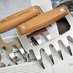 Puede incluir: Un par de garras de barbacoa de acero inoxidable con mangos de madera. Los mangos están grabados con las palabras "Barbecue partner".