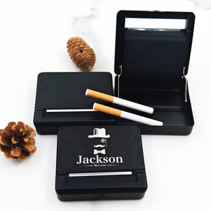 May include: Black rolling box with a hinged lid, featuring a white graphic of a hat, mustache, and bow tie, with the name "Jackson" and "Best man" printed below. Two cigarettes rest on the open box. A pine cone is on the side.