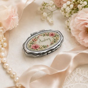 May include: An oval silver-toned metal pill box featuring a hinged lid with a decorative floral border in shades of pink and green. The center of the lid displays the name Audrey in elegant cursive script against a cream-colored background.